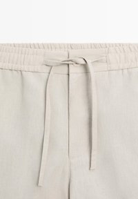 Light beige linen pants with elastic waistband and front tie closure, featuring visible stitching and a single button detail.