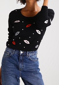 Czarny sweter dziany z kolorowymi wzorami ust w czerwieni, różu i szarości, ozdobiony białymi perłami; okrągły dekolt i długie rękawy.