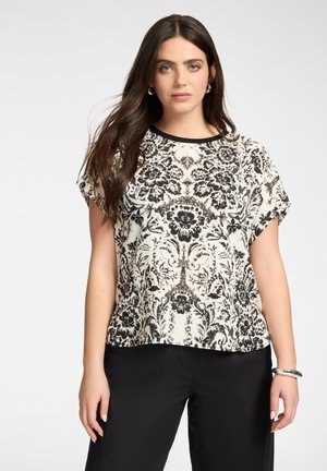 Frau mit langen dunklen Haaren, die ein schwarz-weiß geblümtes kurzärmeliges Oberteil und schwarze Hosen trägt, steht vor einem schlichten Hintergrund.