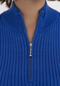 Blå ribbad stickad tröja med dragkedja i kragen. Har en silverfärgad dragkedja och en texturerad yta. Närbild på halsområdet och kragdetaljer.