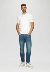 Weißes Poloshirt mit Kragen, blaue Jeans und beige Sneakers. Das Outfit hat eine klassische Passform und minimalen Markenaufdruck auf dem Shirt.
