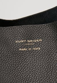 Svart texturerat läder med präglad guldlogotyp "Kurt Geiger London" och "made in Italy." Slät inredning, minimalistisk design.