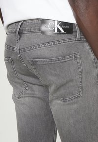 Närbild av en person som bär grå Calvin Klein-jeans som visar bakfickor och varumärkesetikett på midjebandet.
