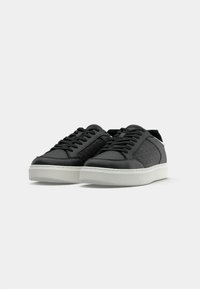 Zwarte sneakers met gestructureerde zijkanten, vlakke witte rubberen zolen en zwarte veters. Het ontwerp heeft een geometrisch patroon aan de zijkanten.