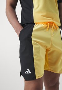 Czarno-żółte spodenki sportowe z elastycznym pasem, charakteryzujące się gładką fakturą i białym logo adidas po lewej stronie.