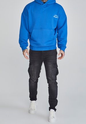 Blauer Kapuzenpullover mit Kängurutasche, kombiniert mit schwarzen Cargo-Jeans mit seitlichen Taschen und weißen Turnschuhen. Glatte Stoffoberfläche.