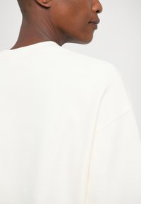 Felpa bianca con una vestibilità rilassata, texture morbida, colletto a girocollo e spalle scese, che presenta un design semplice e fluido.