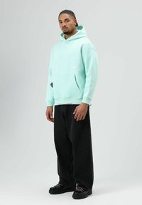 Mintgroene hoodie met kangoeroezak, lange mouwen en geribbelde manchetten, gecombineerd met zwarte wijde jeans en zwarte veterschoenen.