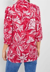 Femme portant une chemise rouge à boutons avec des motifs floraux blancs et des manches trois-quarts, associée à un jean bleu, vue de l'arrière.