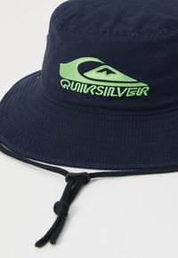 Námornícka modrá vedro klobúk vyrobený z textilu, s zeleným logom Quiksilver. Obsahuje šnúrku na úpravu a textúrované povrchové detaily.