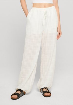 Femme portant un pantalon blanc à carreaux amples avec des poignets élastiques et des sandales noires, debout devant un fond clair uni.