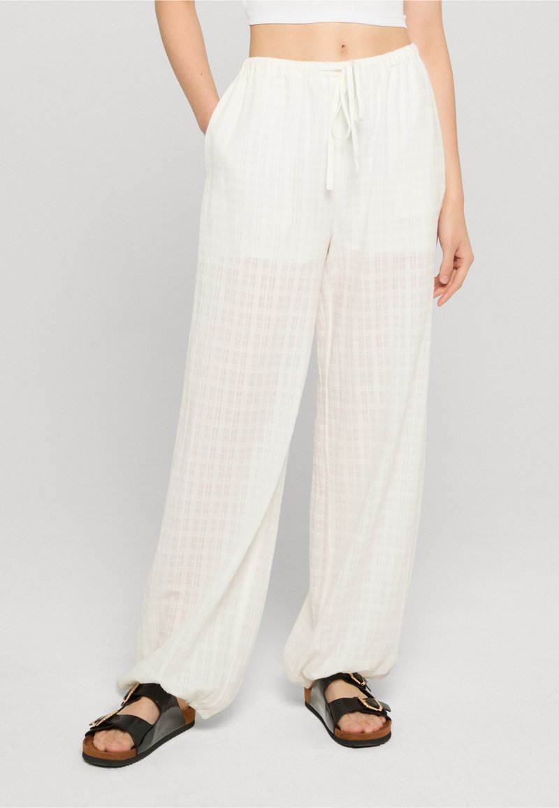 Femme portant un pantalon blanc à carreaux amples avec des poignets élastiques et des sandales noires, debout devant un fond clair uni.