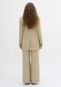 Beige skräddarsydd kostym med en lång kavaj som har en sprund bak, i kombination med vida byxor. Slät textur och strukturerad design.