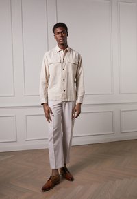 Chemise boutonnière beige clair avec deux poches poitrine, associée à un pantalon gris clair et des chaussures en cuir marron, le tout sur un fond minimaliste.
