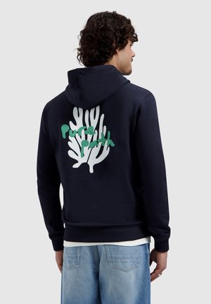 Mann mit lockigem Haar trägt einen marineblauen Hoodie mit weißem Korallenmotiv und grünem Text auf der Rückseite, kombiniert mit hellblauen Jeans.