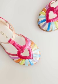 Többszínű szandálok bőr pántokkal, szív alakú díszítéssel. A pántok rózsaszín, sárga, kék és lila árnyalatúak, világos talpon.