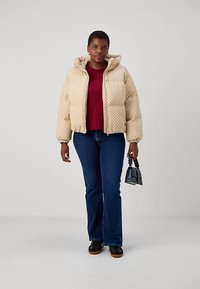 Ženska oblečena v bež prešito kapuco, rdečo majico, modre kavbojke, črne čevlje, drži majhno črno torbico s zvitim ročajem, stoji na enobarvni ozadju.