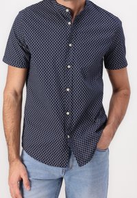 Camisa de manga corta azul marino con pequeños motivos blancos, con cuello mao y tela ligera. Se pueden ver los botones delanteros.