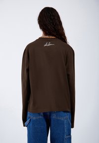 Braunes Langarmshirt mit rundem Halsausschnitt und einem weißen Signaturdetail auf dem oberen Rücken. Kombiniert mit blauen Denim-Cargo-Hosen.