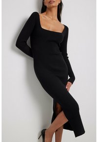 Svart ribbad stickad klänning med långa ärmar, fyrkantig halsringning och sidoslits, figurnära silhuett, mjuk textur, bärs med svarta högklackade skor.