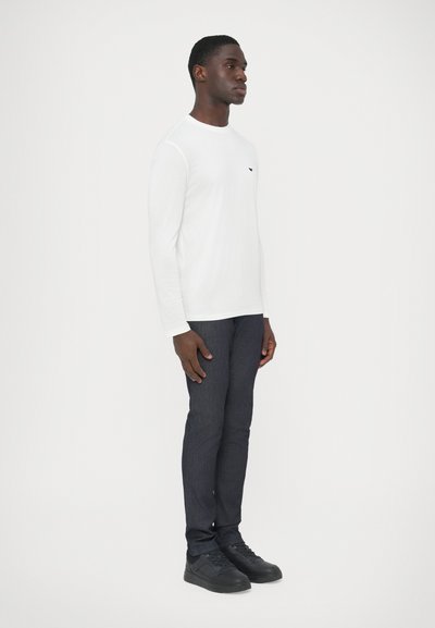 Λευκό πουκάμισο με μακριά μανίκια και στρογγυλή λαιμόκοψη, συνδυασμένο με σκούρο σμιλεμένο τζιν και μαύρα αθλητικά παπούτσια, με ελάχιστες λεπτομέρειες.