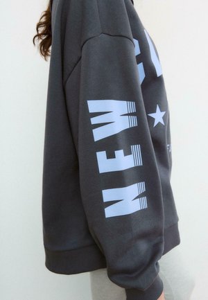 Personne portant un sweat-shirt gris foncé avec un grand texte "NEW" bleu clair sur la manche et un graphique d'étoile sur la poitrine, debout de profil.