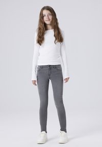 Bela srajca z dolgimi rokavi v kombinaciji s sivimi ozkimi jeans. Jeans imajo pet žepov in so oblikovani z rahlim pranjem. Obutev so bele superge.