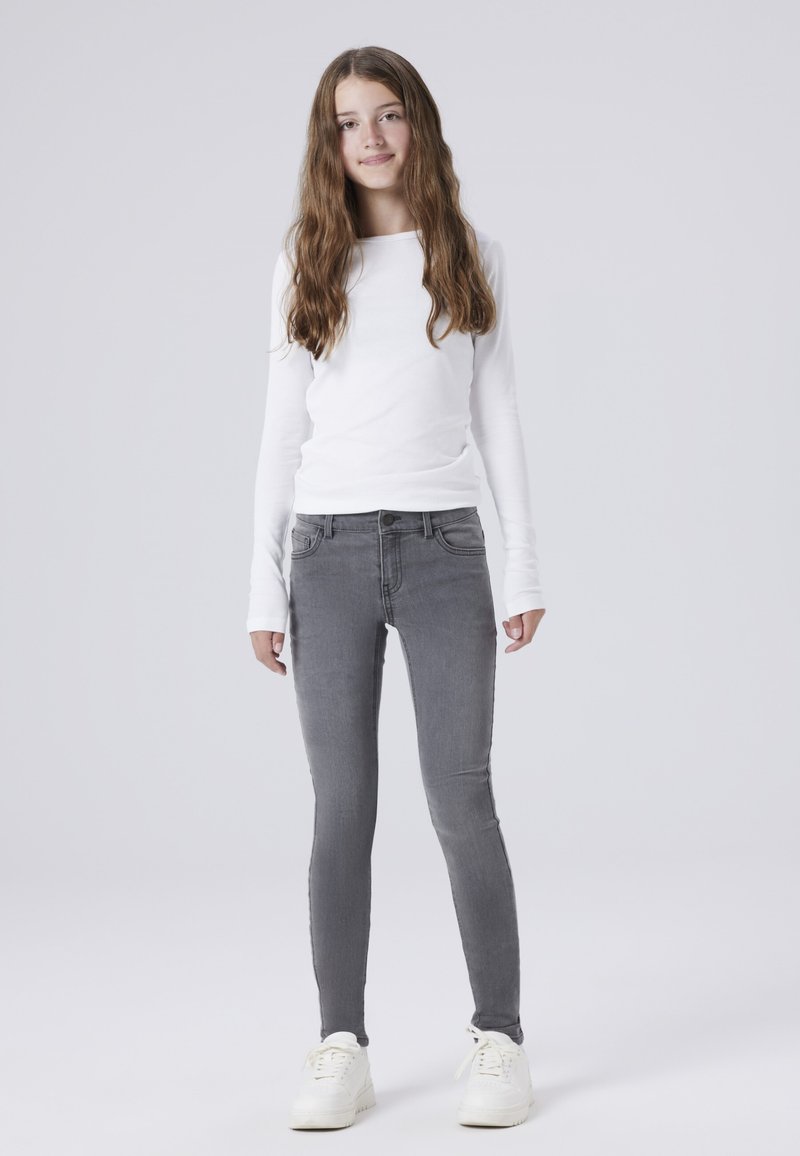 Bílá košile s dlouhým rukávem spárovaná se šedými skinny džíny. Džíny mají pět kapes a jsou zpracovány s jemným omytím. Obuv tvoří bílé tenisky.