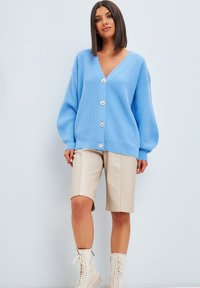 Ljusblå stickad cardigan med djup V-hals, prydd med fem stora knappar, överdimensionerade ärmar, kombinerad med beige shorts och vita botten.