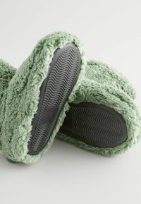 Grüne Plüschschuhe mit einem weichen, pelzigen Äußeren und schwarzem Gummisohle, die ein welliges Profilmuster für besseren Halt aufweist. Größenschild sichtbar.