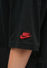 Crna pamučna majica s kratkim rukavima, s crvenim izvezenim Nike logotipom na lijevom rukavu. Glatka tekstura, ležerni dizajn.