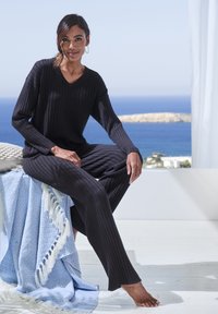 Schwarzes Rippstrick-Loungewear-Set mit langen Ärmeln und weitem Bein. Das Modell sitzt auf einer hellblauen gemusterten Decke mit weißen Quasten.