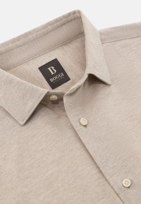 Beżowa, teksturowana koszula z klasycznym kołnierzykiem, z metką BOGGI i dwoma okrągłymi guzikami. Tkanina ma delikatny wzór.