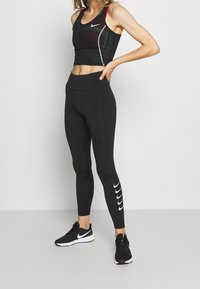 Czarny sportowy crop top z czerwonymi akcentami, zestawiony z czarnymi legginsami z białym wzorem logo Nike po boku oraz czarnymi butami sportowymi.