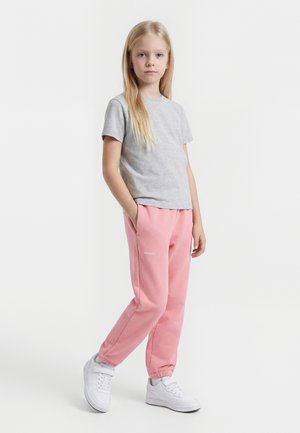 Ung pige står med hænderne i lommerne iført en grå t-shirt, lyserøde joggingbukser og hvide sneakers mod en hvid baggrund.