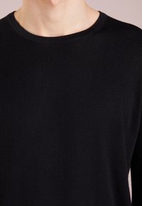Primo piano di una persona che indossa un semplice maglione nero a girocollo, con il collo e la parte superiore del torace visibili su uno sfondo neutro.