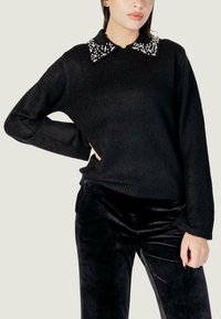 Suéter negro de punto con un cuello adornado con perlas, con mangas largas y un corte holgado, combinado con pantalones de terciopelo negros.