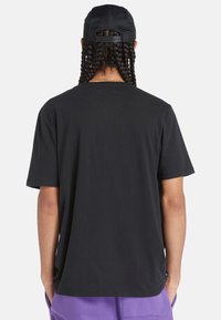 Schwarzes T-Shirt mit kurzen Ärmeln, entspannter Passform, Rundhalsausschnitt und glatter Textur, von hinten betrachtet. Mit minimalem Branding am Saum.