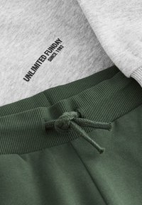 Grauer Pullover mit dem Schriftzug „UNLIMITED FUNDAY SINCE 1982“; grüne Jogginghose mit geripptem Bund und Kordelzugverschluss.