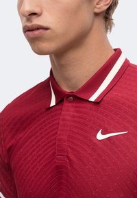Jeune personne portant un polo rouge Nike avec des bandes blanches sur le col et un logo blanc Nike sur la poitrine.