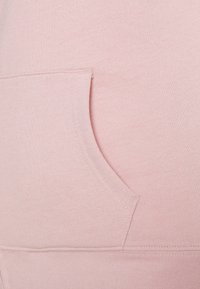 Tessuto di felpa rosa chiaro con una finitura liscia e un dettaglio sulla tasca laterale. Include cuciture lungo i bordi e l'orlo.