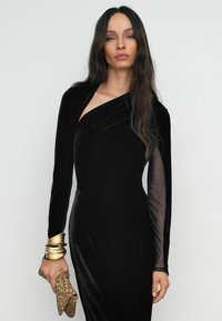 Melnas samta asimetriskas kleitas ar vienu garu punktiņu acs piedurkni. Modelis tur rakstainu somiņu un valkā zelta rokassprādzi.