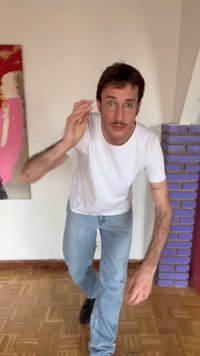 Muž s okuliarmi, v bielom tričku a džínsach sa nakláňa dopredu s pozdvihnutou rukou v miestnosti s abstraktným obrazom a fialovým tehlovým stĺpom.