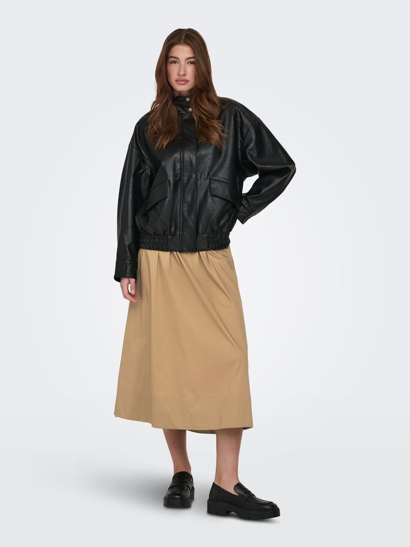 Femme portant une veste en cuir noire, une jupe midi beige et des mocassins noirs, debout devant un fond clair uni.