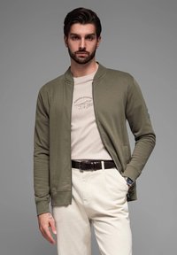 Giacca bomber khaki con zip sopra una maglietta beige con stampa grafica, abbinata a pantaloni crema e una cintura marrone, con un orologio al polso.