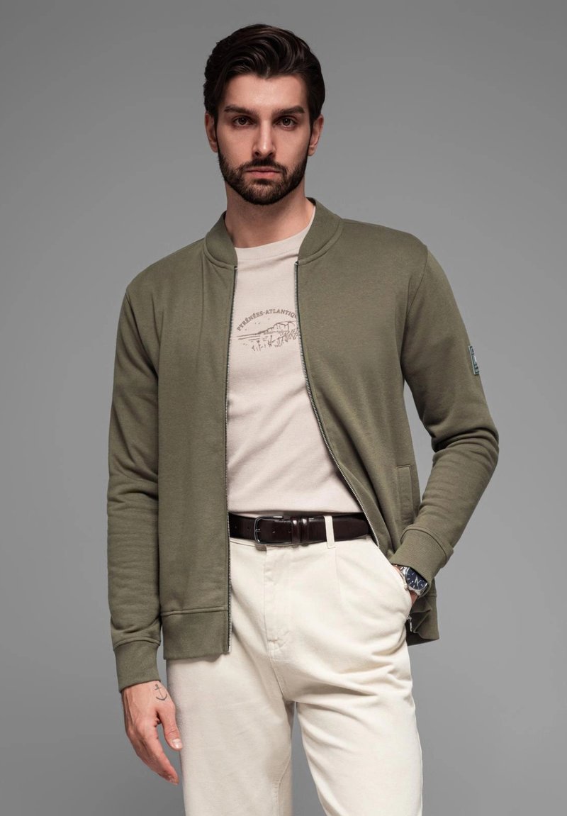 Giacca bomber khaki con zip sopra una maglietta beige con stampa grafica, abbinata a pantaloni crema e una cintura marrone, con un orologio al polso.