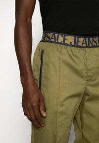 Left hand rests by side wearing olive green pants with black zipper pocket and elastic waistband labeled "Versace Jeans."