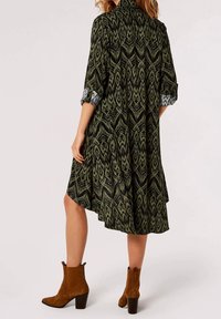 Grün gemustertes Kleid mit einem Schalkragen und dreiviertellangen Ärmeln, ausgestattet mit einem asymmetrischen Saum. Kombiniert mit braunen Knöchelstiefeln.