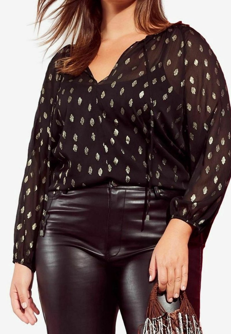 Femme portant un chemisier noir transparent avec des taches dorées et un pantalon en cuir noir, tenant un sac à main marron et blanc orné de perles.