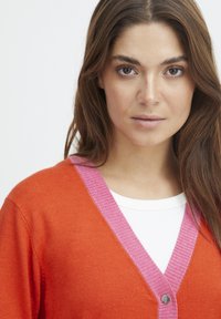Orange stickad cardigan med en ljusrosa ribbad krage och knapplik, lager på en vit topp. Enkelt mönster med kontrasterande färger.